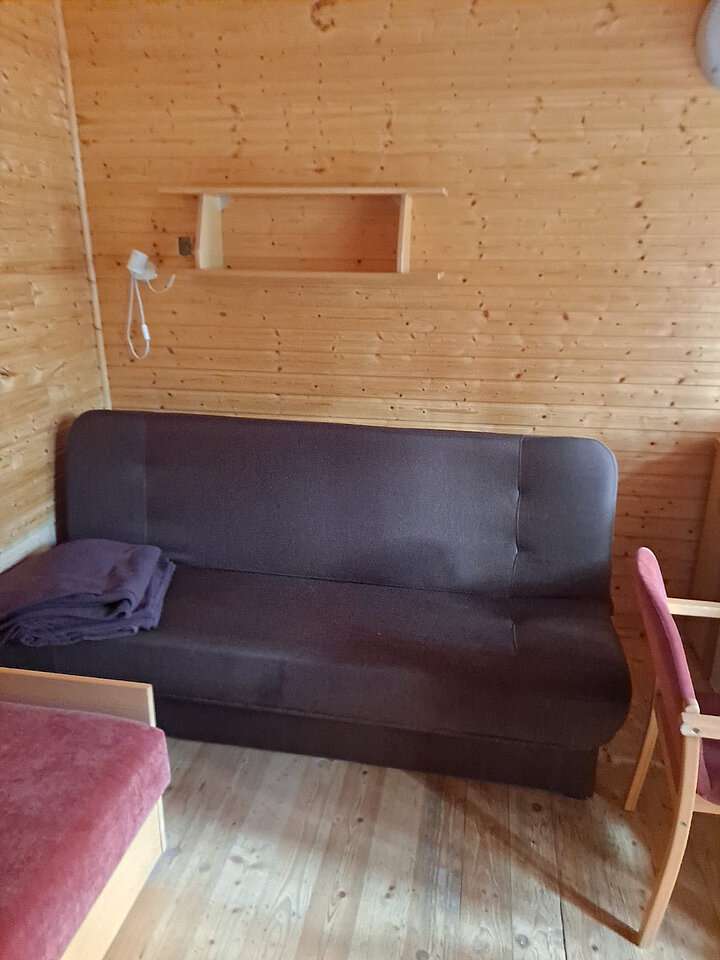 Zdjęcie wnętrza drewnianego pomieszczenia. Widok na kanapę oraz fragment łożka oraz krzesła.