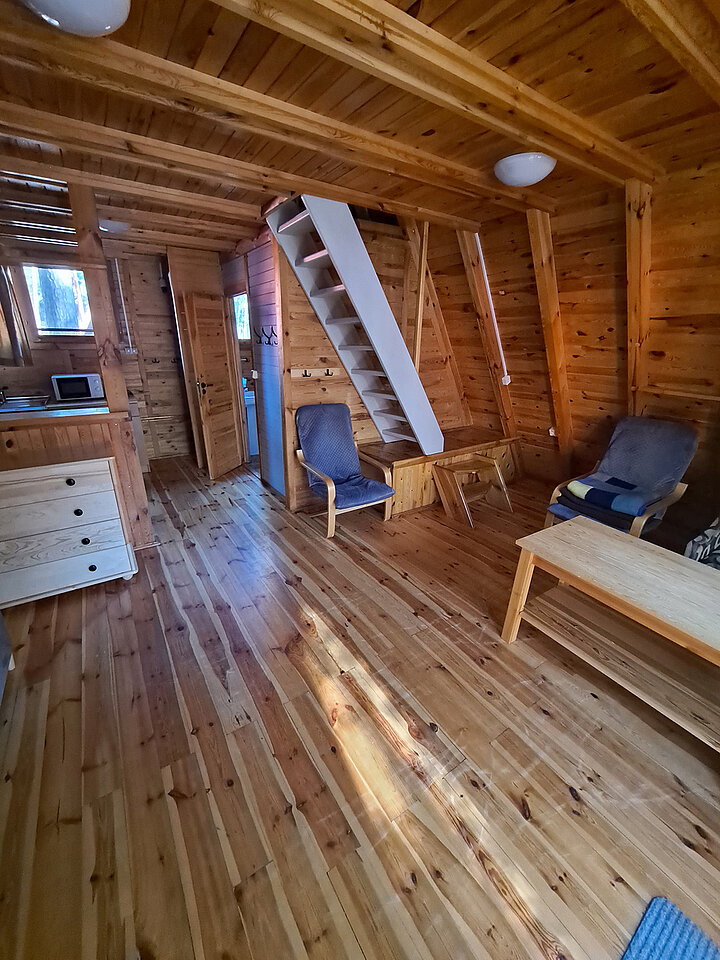 Zdjęcie wnętrza domku ze spadzistym dachem. Widać drewnianą ławę, dwa fotele, komodę oraz strone schody na piętro. W tle mały aneks kuchenny.