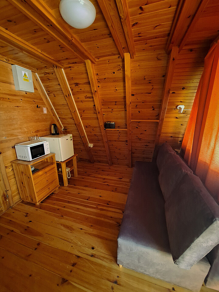 Wnętrze poddasza drewnianego domku turystycznego z mocno spadzistym dachem. Widać rozkłądaną kanapę, drewnianą szafkę z kuchenką mikrofalową oraz niewielką lodówką po jej prawej stronie.