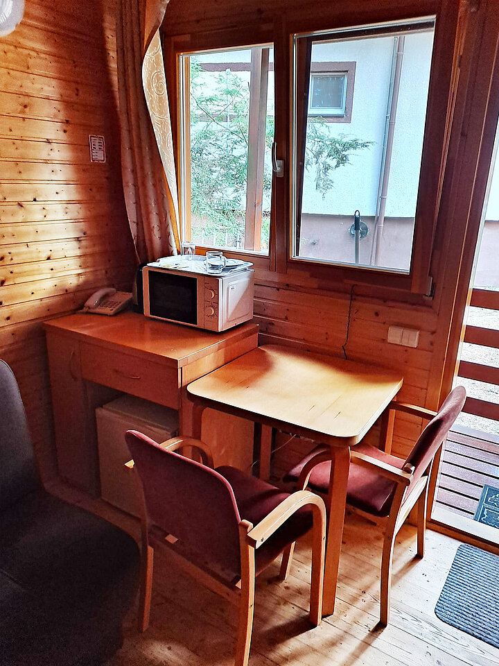 Zdjęcie wnętrza drewnianego pomieszczenia. Widok na przeszkoną ścianę. Przed nią stolik z dwoma krzesłami oraz mała szafka z lodówką i kuchenką mikrofalową.