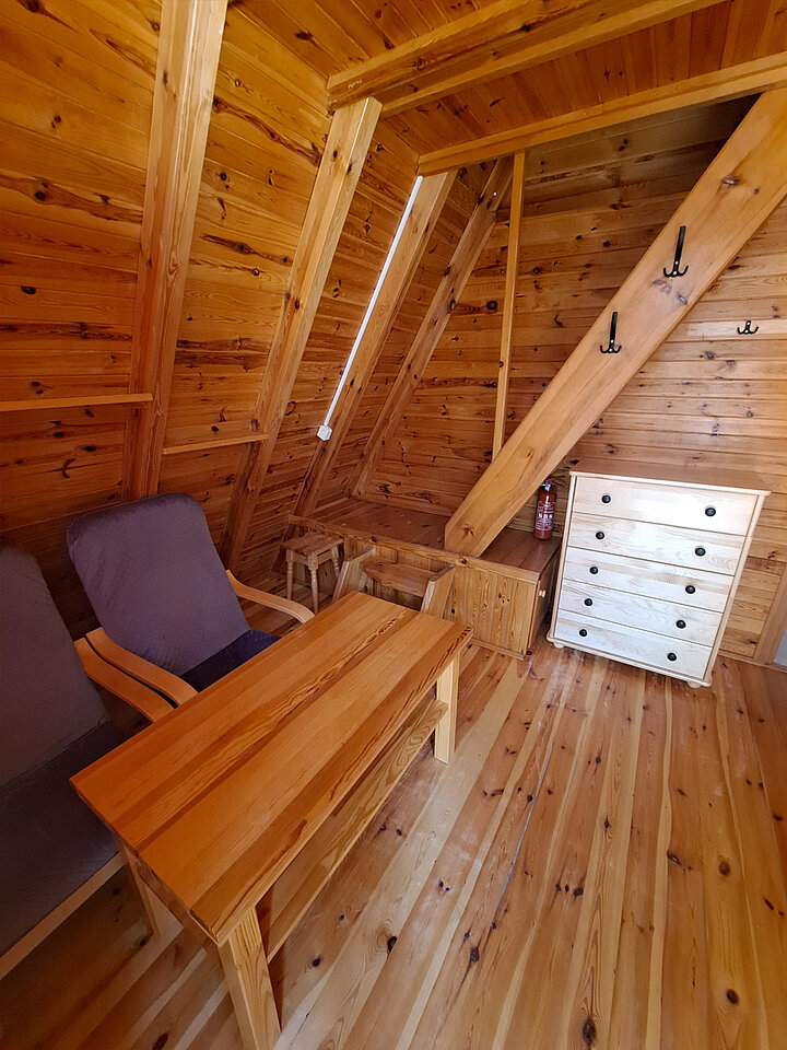 Wnętrze dreewnianego domkui turystycznego z mocno spadzistym dachem. Widać sosnowy stółi dwa fotele. W tle schody na piętro.